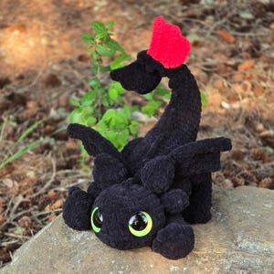 Pode incluir: Um drag&atilde;o preto feito &agrave; m&atilde;o em croch&ecirc; com uma barbatana vermelha na cabe&ccedil;a. O drag&atilde;o tem olhos grandes e redondos com detalhes em verde e amarelo. Est&aacute; sentado em uma rocha cinza, com um fundo natural de vegeta&ccedil;&atilde;o e terra marrom.