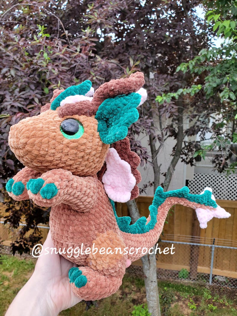 May include: A brown crocheted dragon with green and pink accents. The dragon has large green eyes and a long tail. The dragon is holding its paws up in the air. The text "@snugglebeanscrochet" is visible in the bottom left corner of the image.