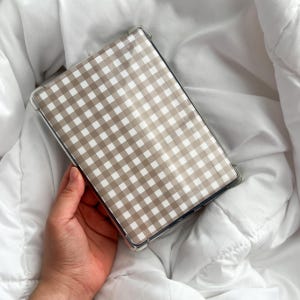 K&ouml;nnte beinhalten: Eine transparente Schutzh&uuml;lle f&uuml;r ein Tablet mit einem beige-wei&szlig;en Vichy-Muster. Die rechteckige H&uuml;lle bietet Schutz. Der Hintergrund ist ein wei&szlig;es, strukturiertes Gewebe.