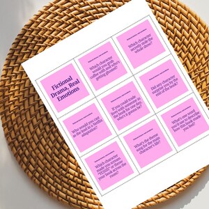 Puede incluir: Una hoja de papel blanca con cuadrados rosas que contienen preguntas sobre libros, titulada "Fictional Drama, Real Emotions". El papel est&aacute; sobre una superficie de mimbre tejido. Las preguntas incluyen "&iquest;Con qu&eacute; personaje tomar&iacute;as caf&eacute;?" y "&iquest;Cu&aacute;l es la canci&oacute;n principal?"