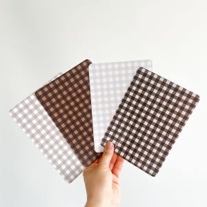 K&ouml;nnte beinhalten: Eine Hand h&auml;lt vier rechteckige Tischsets mit Vichy-Muster. Die Tischsets variieren in Braun- und Beiget&ouml;nen. Der Hintergrund ist schlicht wei&szlig;.