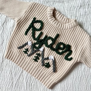 Könnte beinhalten: Ein cremefarbener Strickpullover mit einem grün-weißen Stickmuster, auf dem "Rider" mit einer Bergszene steht.