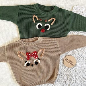 May include: Two knitted sweaters with embroidered reindeer faces. The sweater on the left is green and the sweater on the right is brown. The reindeer on the green sweater has a red nose and the reindeer on the brown sweater has a red bow.
