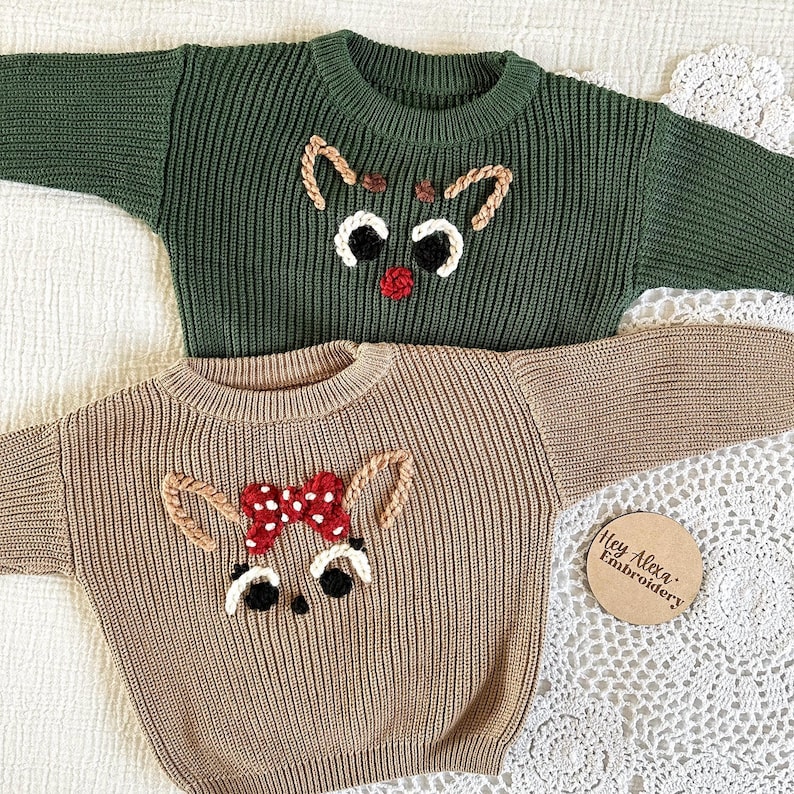 May include: Two knitted sweaters with embroidered reindeer faces. The sweater on the left is green and the sweater on the right is brown. The reindeer on the green sweater has a red nose and the reindeer on the brown sweater has a red bow.