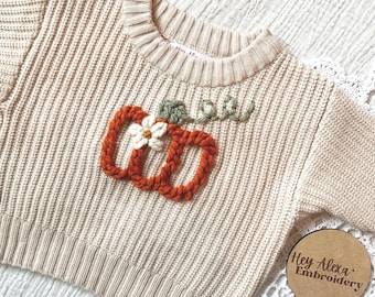May include: A cream-colored knitted sweater with a hand-stitched pumpkin design in orange and green. The sweater has a ribbed texture and a crew neck. The text "Hey Alexa Embroidery" is on a brown tag.
