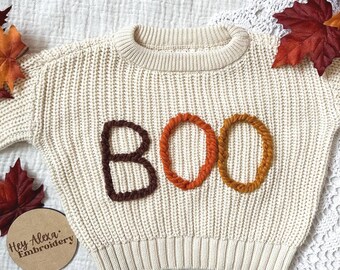 Puede incluir: Un suéter de punto color crema con la palabra "BOO" bordada en hilo marrón, naranja y amarillo.