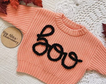 Puede incluir: Un suéter de punto de color melocotón con un "BOO" bordado en negro en la parte delantera. El suéter está hecho de un material suave y cómodo y es perfecto para la temporada de otoño.