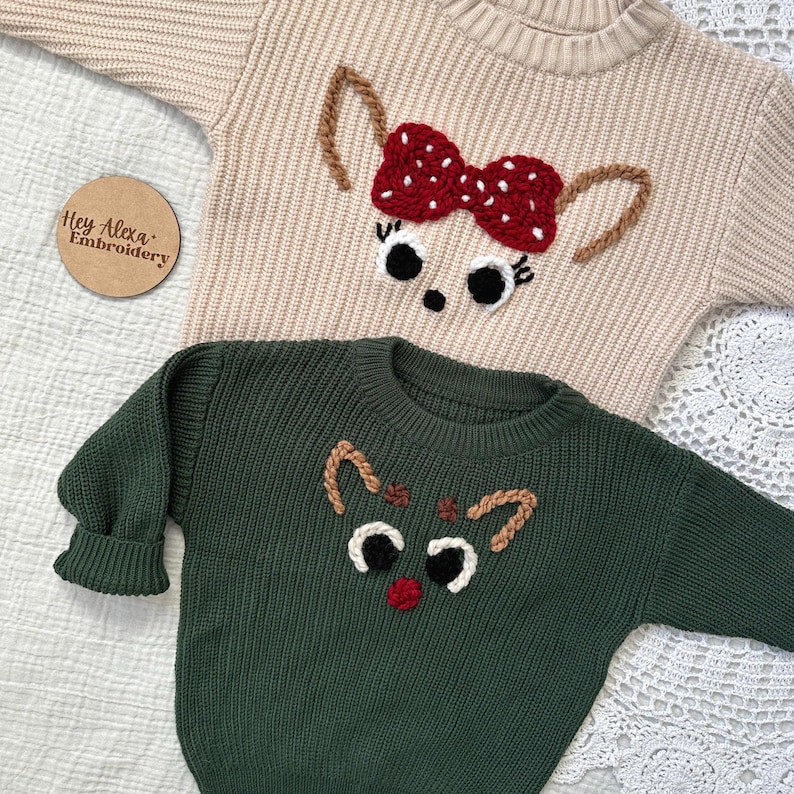 May include: Two knitted sweaters with embroidered reindeer designs. The top sweater is beige with a red bow and the bottom sweater is green with a brown nose.