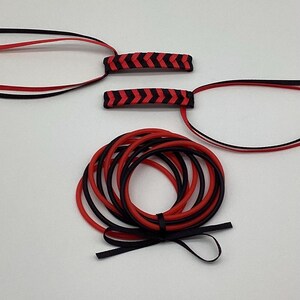 May include: An assortment of black and red cords and accessories. Two small, rectangular pieces with a chevron pattern in red and black are at the top. Below are several coiled cords in red and black, with a black ribbon tied at the bottom.