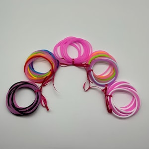 May include: A collection of colorful rubber bracelets, including pink, black, white, and rainbow hues. Each bundle is tied with a pink ribbon. The bracelets are arranged on a white surface.