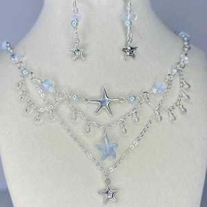 May include: A silver-toned necklace and earring set featuring starfish and clear and blue beads. The necklace has multiple layers with a large starfish pendant and smaller starfish charms. The earrings have a similar design with dangling beads.