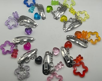 Könnte beinhalten: Sortiment an bunten Haarklammern mit Charms. Jede Klammer ist mit einem Stern-, Herz- oder Bären-Charm sowie einer Kette mit zusätzlichen Charms verziert. Die Charms sind in verschiedenen Farben, darunter Pink, Blau, Grün, Rot und Gelb.