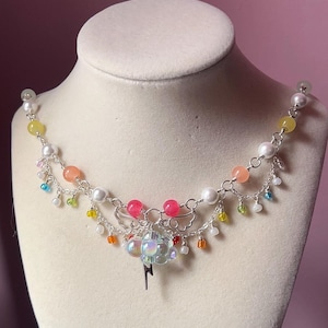 May include: A silver-toned necklace with multiple strands, adorned with colorful beads, pearls, and a cloud-shaped charm with a lightning bolt pendant. The necklace is displayed on a mannequin bust.