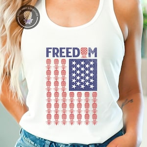 May include: White tank top with a patriotic design. The design features the word "FREEDOM" above a flag made of red pineapples and a blue square with white stars. The tank top is paired with denim shorts.