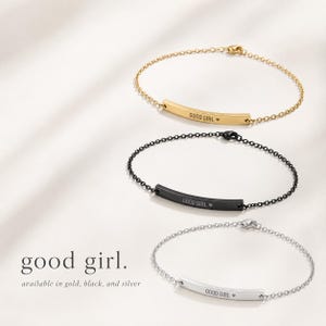 May include: Three delicate chain bracelets, each featuring a curved metal bar engraved with the text GOOD GIRL and a small heart symbol. The set includes one gold-tone, one black, and one silver-tone bracelet, all with matching metal clasps.