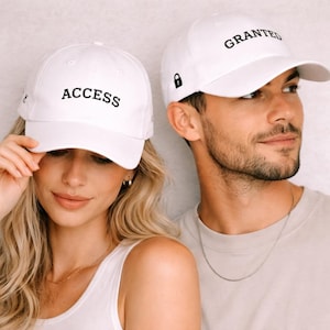 May include: Two white baseball caps. One cap has the word "ACCESS" embroidered in black, the other has "GRANTED". Both caps have a curved brim and a black lock symbol. The caps are worn by two people.
