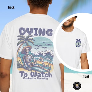 May include: White t-shirt with a graphic on the back featuring a skeleton in a beach chair under a palm tree, with the words "DYING To Watch" and "Cucked in Paradise". A smaller palm tree graphic is on the front.