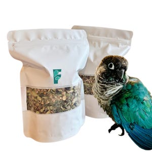 May include: Two white stand-up pouches with clear windows displaying a dry bird food mix. Each bag features a green logo with a bird and hedgehog icon. A green-cheeked conure with dark brown, green, and blue feathers stands beside the bags.