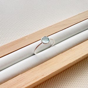 May include: A silver ring with a light blue gemstone set in a twisted band. The ring is displayed in a wooden ring box.