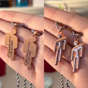 May include: Two copper-colored pendant necklaces. One pendant reads "BIRDS OF A FEATHER" and the other reads "WE SHOULD STICK TOGETHER". The pendants are shaped like a person. The necklaces have a silver chain.