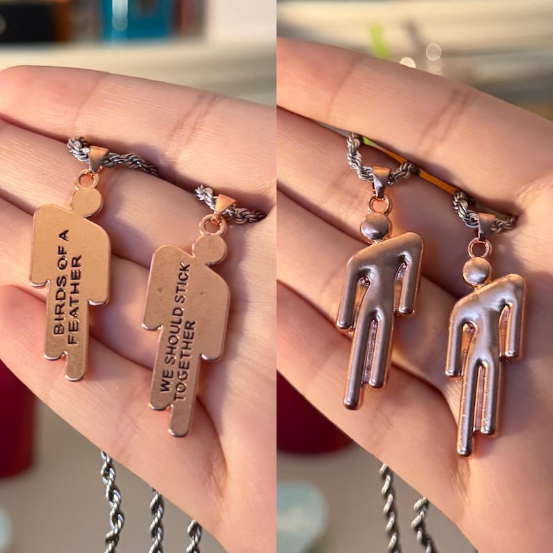 May include: Two copper-colored pendant necklaces. One pendant reads "BIRDS OF A FEATHER" and the other reads "WE SHOULD STICK TOGETHER". The pendants are shaped like a person. The necklaces have a silver chain.