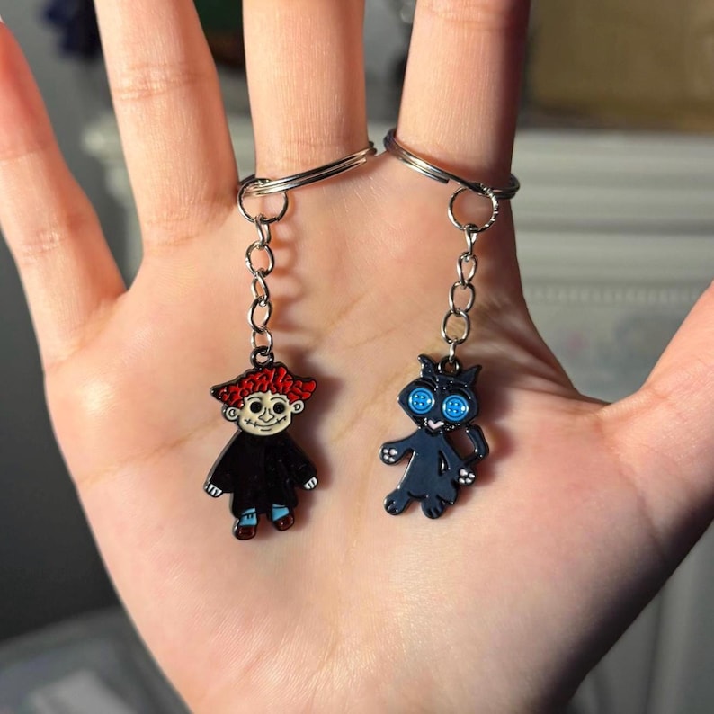 May include: Two keychains with character charms. One keychain features a charm of a character with red hair, a black coat, and blue shoes. The other keychain has a black cat charm with blue button eyes. Both charms are attached to silver keyrings.