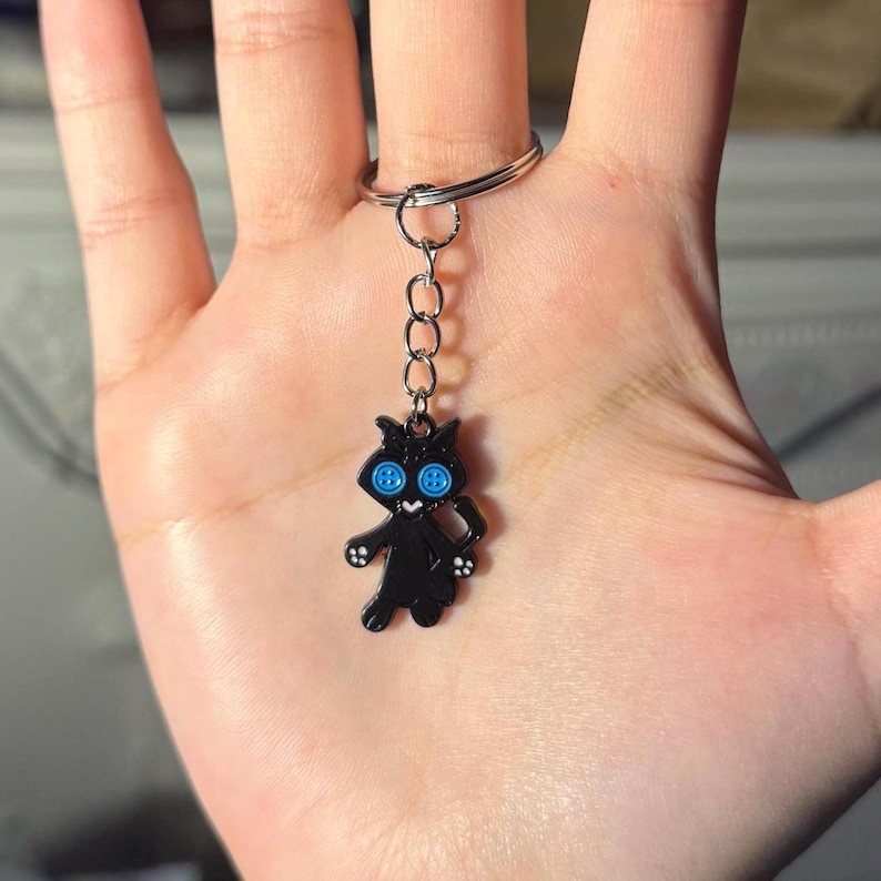 May include: A black cat keychain with blue button eyes and button accents on its paws. The cat charm is attached to a silver chain and keyring. The keychain is a whimsical accessory.