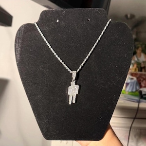 May include: A silver-colored necklace with a pendant in the shape of a person. The pendant and chain are displayed on a black jewelry stand. The pendant is encrusted with small, sparkling stones.