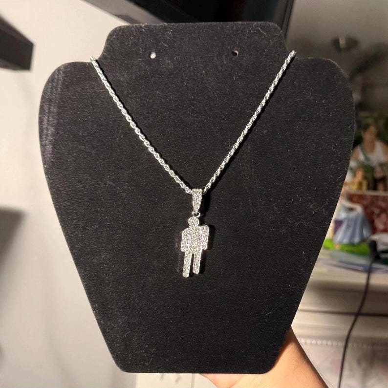 May include: A silver-colored necklace with a pendant in the shape of a person. The pendant and chain are displayed on a black jewelry stand. The pendant is encrusted with small, sparkling stones.