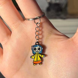 May include: A keychain featuring a cartoon character with blue hair, a yellow coat, and an orange skirt. The character is attached to a silver chain and keyring. The keychain is held up by a hand.