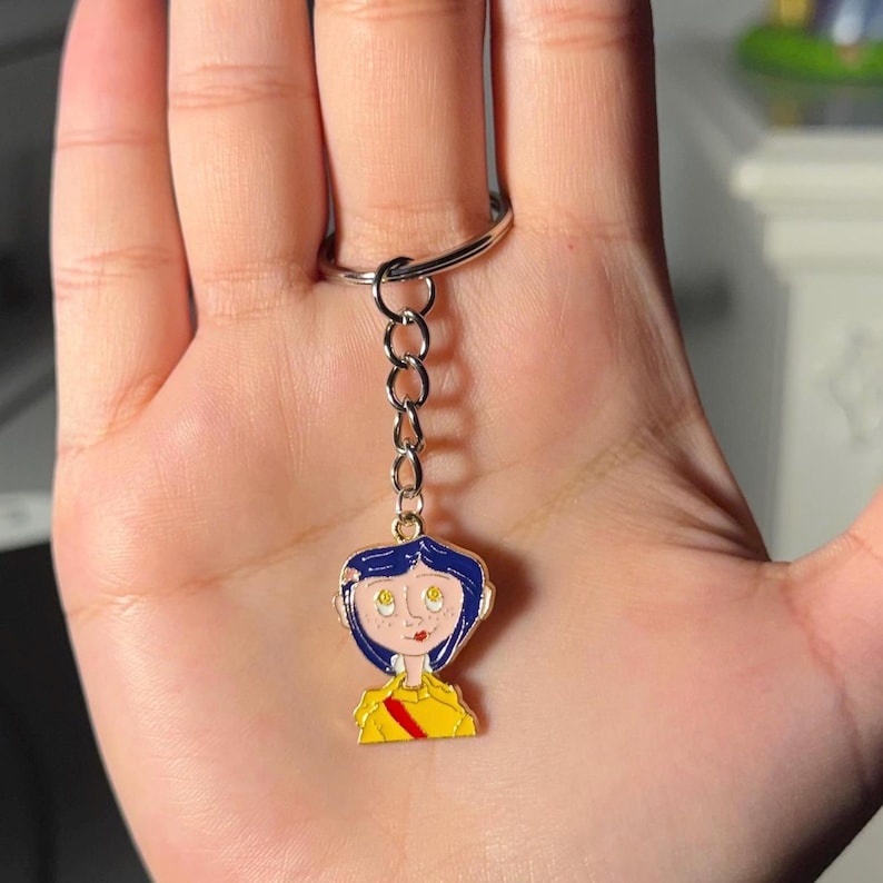 May include: A keychain featuring a cartoon character with blue hair, yellow shirt, and red stripe. The charm is attached to a silver chain and ring. The character has a pale complexion and is smiling.