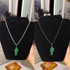 May include: Two necklaces displayed on black stands. One features a silver chain with a green pendant in the shape of a person. The other has a black chain with the same green pendant. The pendants are detailed with small, sparkling stones.