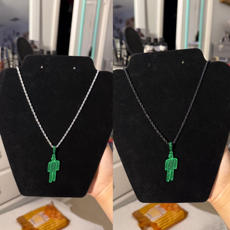 May include: Two necklaces displayed on black stands. One features a silver chain with a green pendant in the shape of a person. The other has a black chain with the same green pendant. The pendants are detailed with small, sparkling stones.