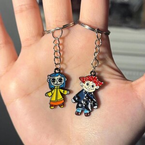May include: Two keychains featuring cartoon characters. One keychain has a character with blue hair, a yellow coat, and orange pants. The other keychain has a character with red hair and a black coat. Both keychains have silver chains and rings.
