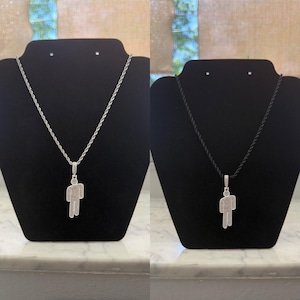 May include: Two necklaces displayed on black stands. One features a silver chain with a pendant in the shape of a person, encrusted with small, sparkling stones. The other has a black cord with a similar pendant.