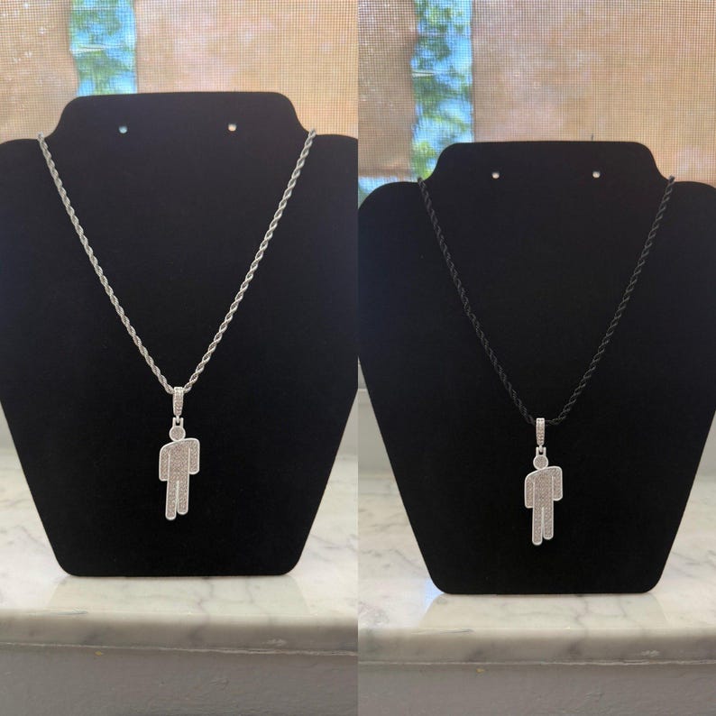May include: Two necklaces displayed on black stands. One features a silver chain with a pendant in the shape of a person, encrusted with small, sparkling stones. The other has a black cord with a similar pendant.