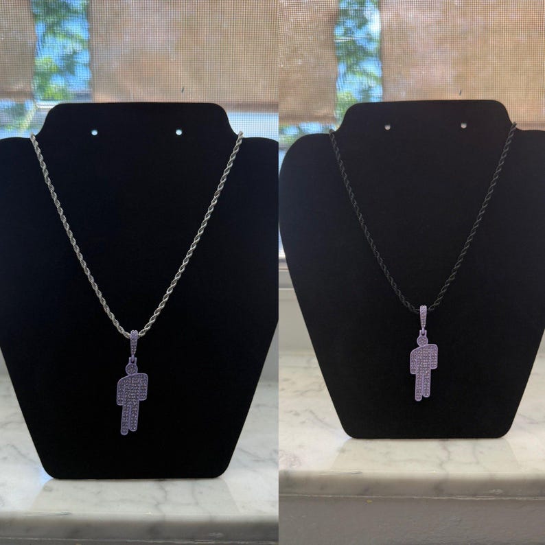 May include: A silver-toned necklace with a pendant in the shape of a person. The pendant is encrusted with small, light purple stones. The necklace is displayed on a black velvet jewelry stand.