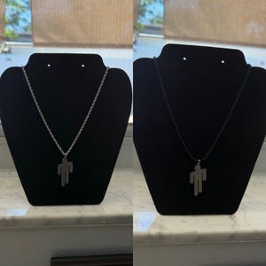 May include: Two necklaces displayed on black stands. One features a silver-colored chain with a pendant in the shape of a person. The other necklace has a black cord and a similar pendant. Both are displayed against a window background.