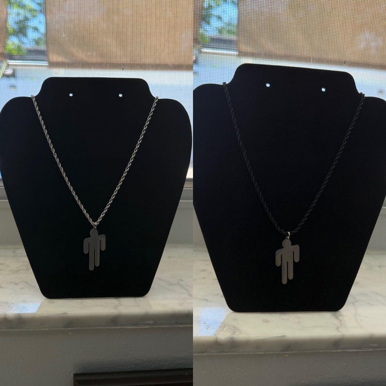 May include: Two necklaces displayed on black stands. One features a silver-colored chain with a pendant in the shape of a person. The other necklace has a black cord and a similar pendant. Both are displayed against a window background.