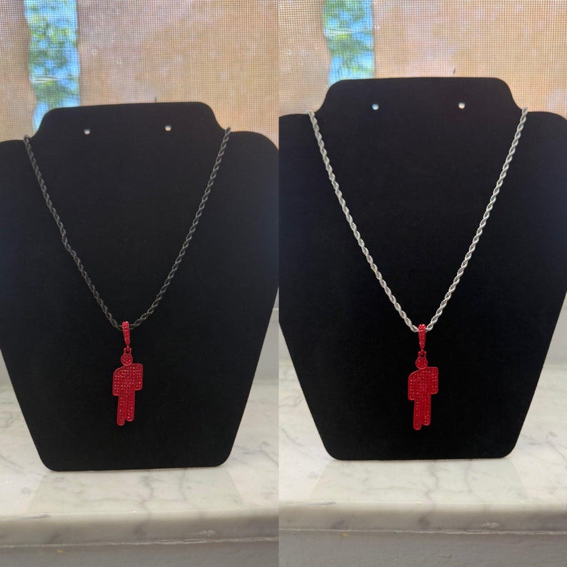 May include: Two necklaces displayed on black stands. Each necklace features a red pendant in the shape of a person. One necklace has a black chain, and the other has a silver chain. The pendants are covered in small, sparkling red stones.