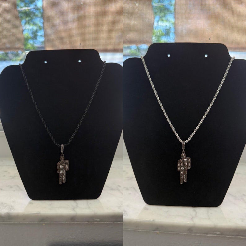 May include: Two necklaces displayed on black stands. One features a black rope chain with a pendant in the shape of a person covered in small, sparkling stones. The other has a silver rope chain with a similar pendant.