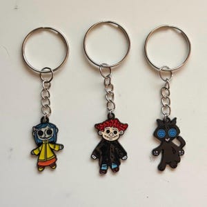 May include: Three keychains featuring characters from a movie. One keychain has a character with blue hair and a yellow coat. Another has a character with red hair and a black coat. The third keychain has a black cat with button eyes.