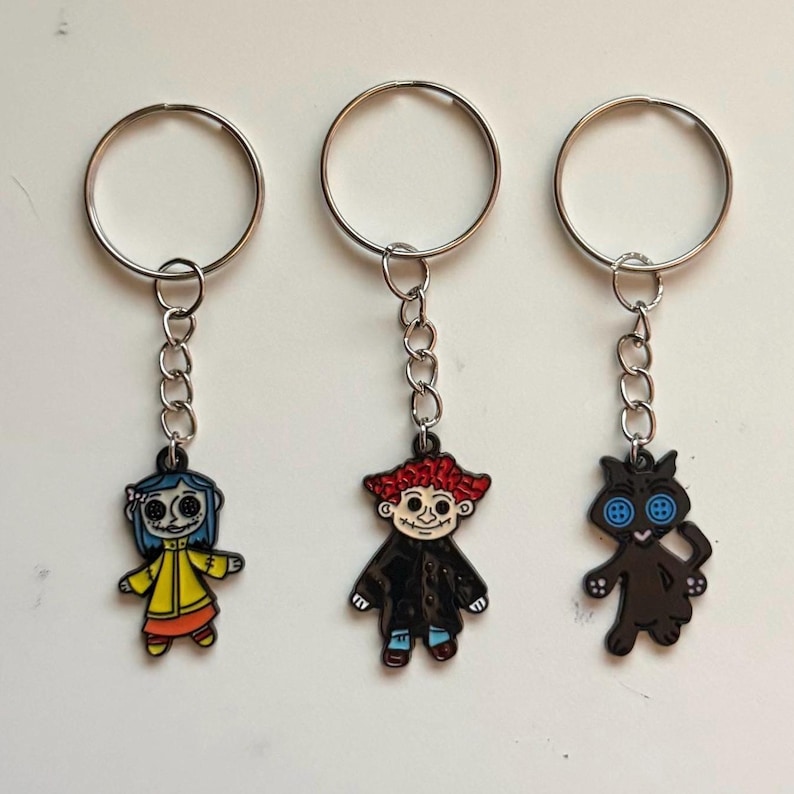 May include: Three keychains featuring characters from a movie. One keychain has a character with blue hair and a yellow coat. Another has a character with red hair and a black coat. The third keychain has a black cat with button eyes.