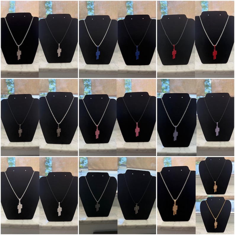 May include: A collection of pendant necklaces featuring a stylized human figure. The pendants come in various colors, including silver, black, blue, red, pink, and gold. Each pendant hangs from a chain or cord, displayed on a black velvet bust.