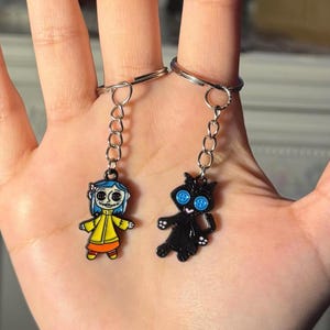 May include: Two keychains featuring cartoon characters. One keychain has a girl in a yellow coat with blue hair. The other keychain has a black cat with blue button eyes. Both keychains are attached to silver keyrings.