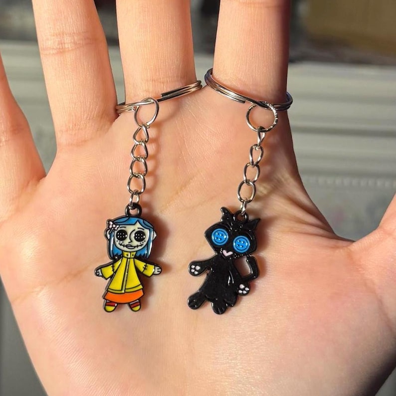 May include: Two keychains featuring cartoon characters. One keychain has a girl in a yellow coat with blue hair. The other keychain has a black cat with blue button eyes. Both keychains are attached to silver keyrings.