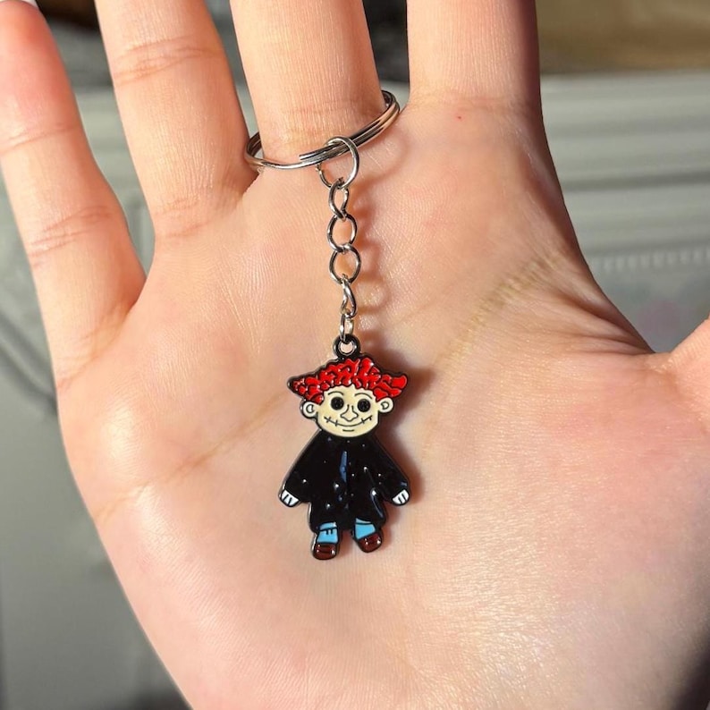 May include: A metal keychain featuring a cartoon figure with red hair, a black coat, and blue pants. The keychain has a silver ring and chain. The figure has a smiling face and is a fun accessory.