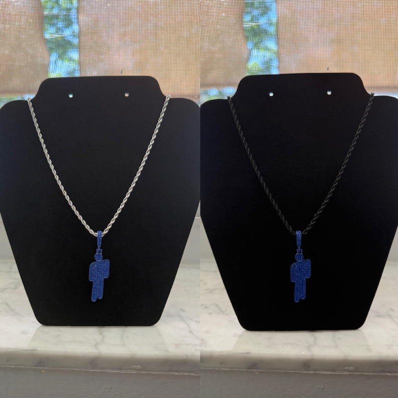 May include: Two necklaces on black display stands. One features a silver chain with a blue pendant in the shape of a person. The other has a black chain with a matching blue pendant. The pendants are covered in small blue stones.