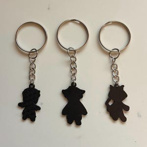 May include: Three black keychains with silver key rings. Each keychain features a different silhouette design: a person, a person in a dress, and a cat. The keychains are connected to the rings with a small chain.
