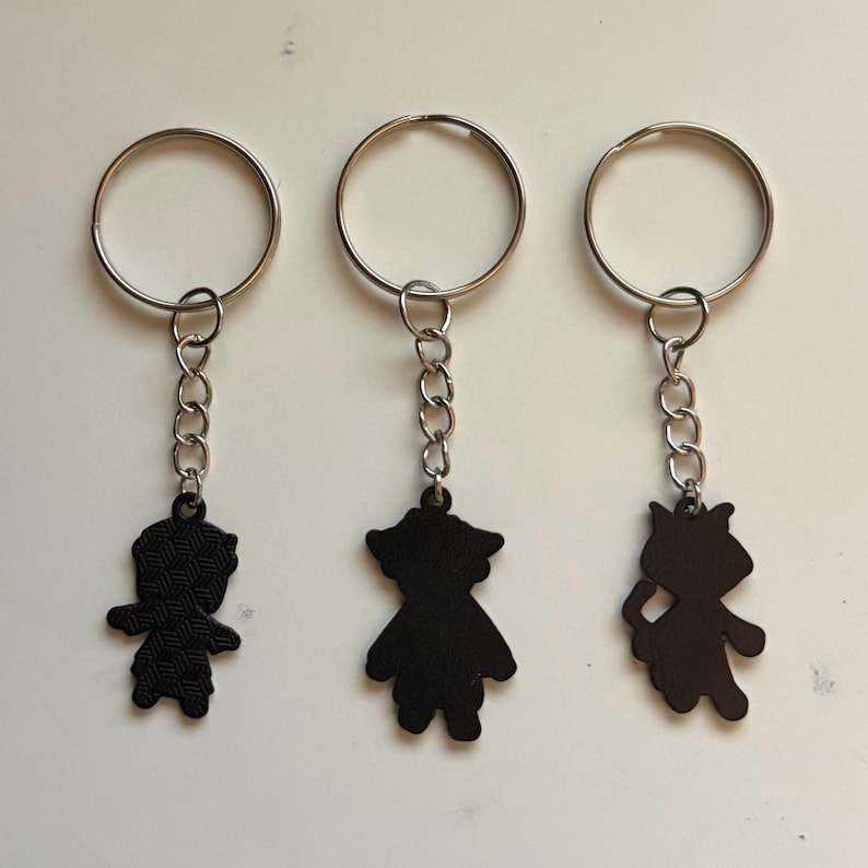 May include: Three black keychains with silver key rings. Each keychain features a different silhouette design: a person, a person in a dress, and a cat. The keychains are connected to the rings with a small chain.